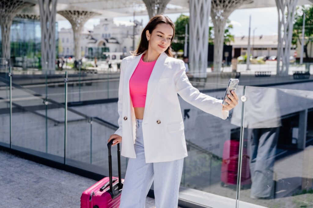 Stylish young woman taking a selfie at the airport while holding a pink suitcase, wearing a white blazer and pink top — chic travel outfit inspiration for modern travelers.
